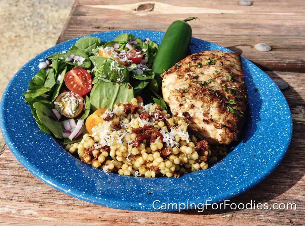 Grilled Dijon Chicken With Garlic And Jalapeño by CampingForFoodies features a blue camping plate filled with a dinner comprised of a golden brown chicken breast with charred grill marks and sprinkled with bright green parsley, on the side is a green dinner salad, a serving of couscous and a whole green jalapeno pepper. The plate is set on a wooden picnic table on a sunny afternoon.