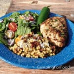 Grilled Dijon Chicken With Garlic And Jalapeño by CampingForFoodies features a blue camping plate filled with a dinner comprised of a golden brown chicken breast with charred grill marks and sprinkled with bright green parsley, on the side is a green dinner salad, a serving of couscous and a whole green jalapeno pepper. The plate is set on a wooden picnic table on a sunny afternoon.