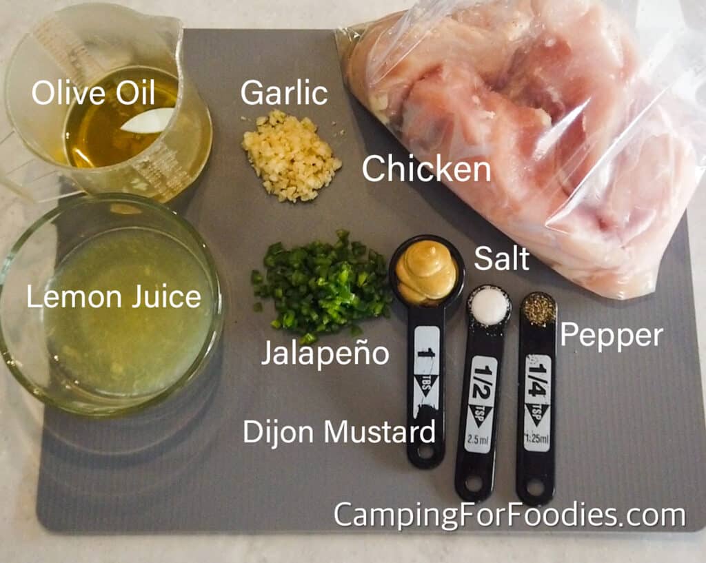 Ingredients for Grilled Dijon Chicken by CampingForFoodies features a gray flexible cutting mat filled with minced yellow-colored garlic, minced green jalapeno pepper, golden olive oil in a measuring cup, fresh-squeezed yellow lemon juice in a bowl, pink chicken breasts in a clear freezer bag and black measuring spoons filled with Dijon mustard, white salt and black pepper.