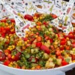 Marinated Corn Salad With Cucumbers And Tomatoes by CampingForFoodies features a bowl filled with a bright colorful salad that includes bright yellow corn kernels, light green diced cucumbers, bright red cherry tomatoes that are sliced in half, chopped red bell peppers and bright green cilantro. The salad glistens from being marinated and tossed in a homemade balsamic vinegar dressing. The bowl’s pattern features retro camping scenes. A matching salad spoon set is placed in the salad and ready for serving.