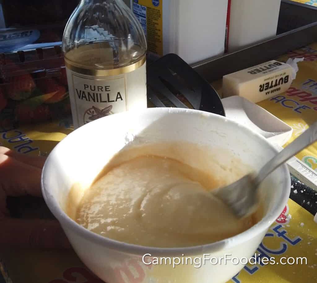 Prepare icing by CampingForFoodies features a small bowl filled with cream cheese icing. A fork is being used to mix the ingredients together. In the background is a bottle of pure vanilla, a stick of butter, a container of milk and a container of whole strawberries.