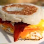Camping Breakfast Sandwiches by CampingForFoodies features an egg, bacon and cheese sandwich on an English muffin. The slices of thick crispy bacon are overflowing the sandwich, the meat is draped over the fried egg and melted yellow cheddar cheese.