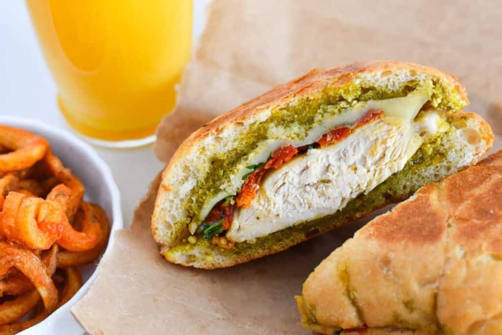 Pesto Chicken Sandwich features a chicken sandwich cut in half, a bowl of curly fries and orange juice in the background.