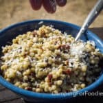 Pesto Couscous Recipe by CampingForFoodies features a blue camping bowl filled with fluffy small round pasta balls, chopped deep red sun dried tomatoes, green capers and white shredded Parmesan cheese. The bowl is set on a picnic table on a sunny day. A spoon is inserted into the dish and a hand is sprinkling cheese on the couscous.