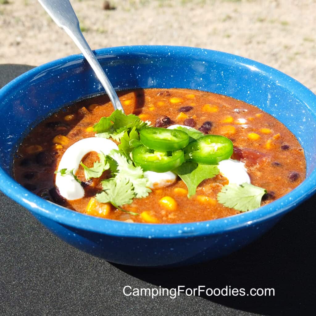 Black Bean And Corn Soup With Chorizo by CampingForFoodies features a blue camping bowl filled with soup with black beans, corn kernels, tomatoes and topped with a drizzle of sour cream, sliced jalapenos and fresh cilantro leaves.