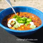 Black Bean And Corn Soup With Chorizo by CampingForFoodies features a blue camping bowl filled with soup with black beans, corn kernels, tomatoes and topped with a drizzle of sour cream, sliced jalapenos and fresh cilantro leaves.