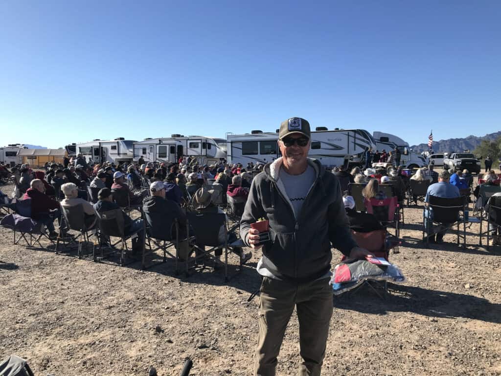 My brother-in-law just won the Grand Design Quartzsite RV rally chili contest features a guy holding a trophy and prizes with a group of people and RVs in the background.