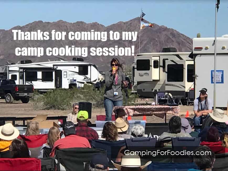 Thanks for coming to my camp cooking session by CampingForFoodies features Kim Hanna on a stage speaking to a crowd. RVs and mountains are in the background.