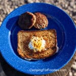 Hobo Eggs by CampingForFoodies features a blue camping plate with an egg that has been fried in the center of the slice of bread that has a hole cut in the center. Two breakfast sausage patties are next to the toasted bread and egg.