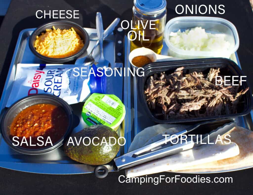 Shredded Beef Burrito Ingredients by CampingForFoodies features a table filled with ingredients including shredded cheese, olive oil, onions, sour cream, taco seasoning, cooked shredded beef, salsa, guacamole, avocado and tortillas. Spoons, a knife and cooking tongs are with the food.