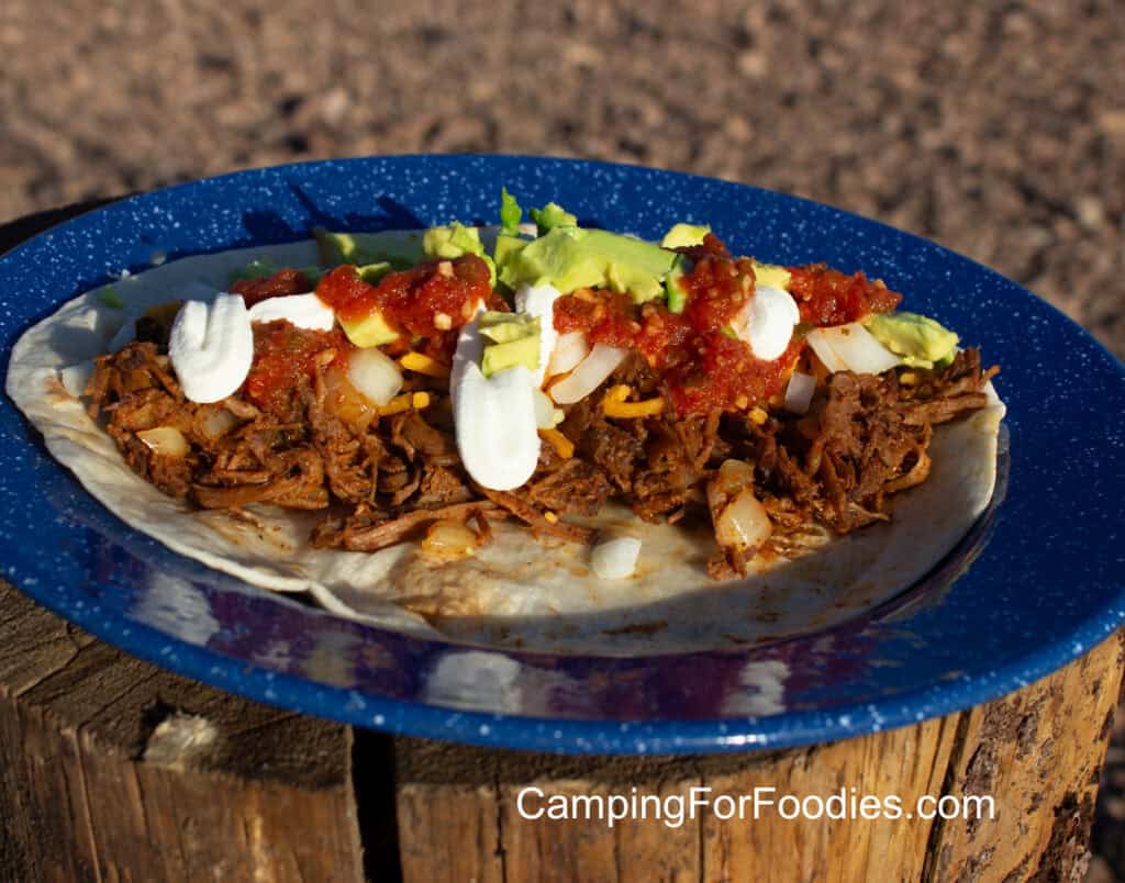 Shredded Beef Burritos by CampingForFoodies features a blue camping plate filled with a tortilla topped with shredded beef, shredded cheese, onions, salsa, cubed avocado and drizzled with sour cream.
