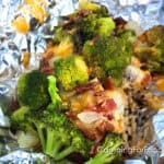 Cheesy Campfire Chicken Foil Packets by CampingForFoodies features a meal on a piece of aluminum foil including chicken, bright green broccoli, bacon and melted cheese.