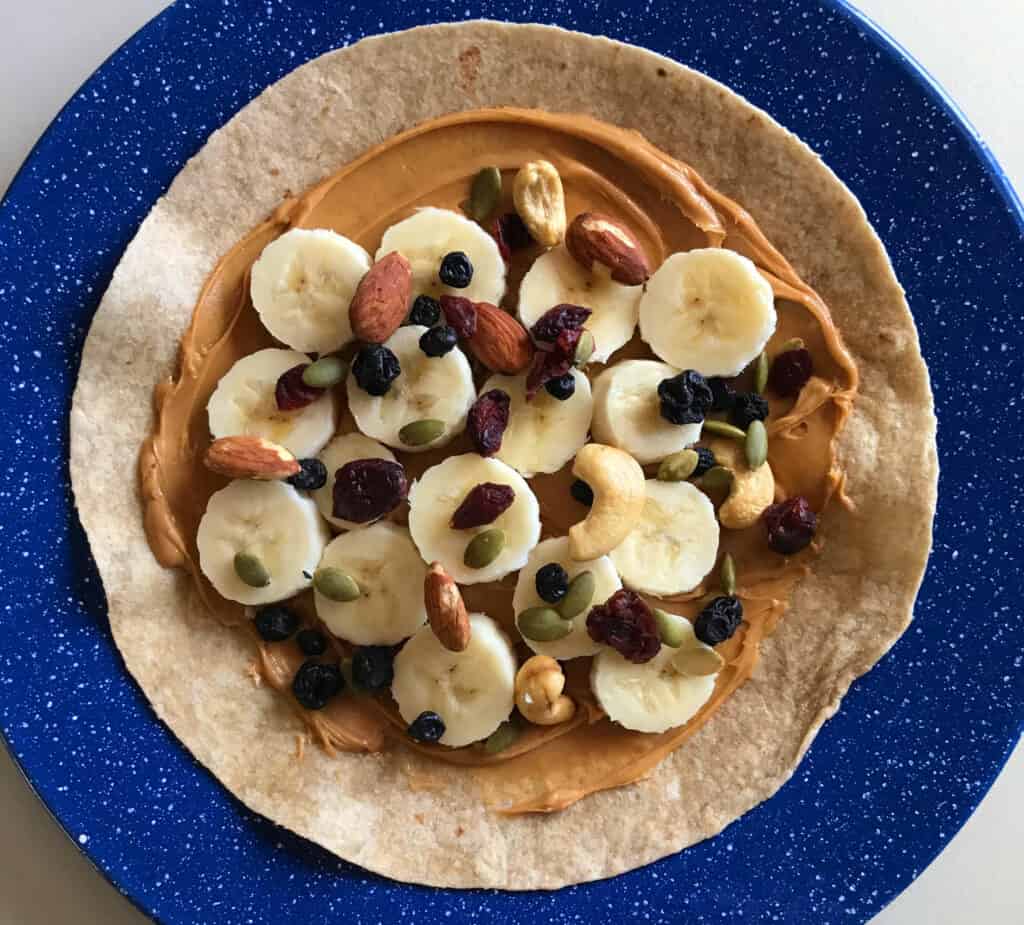 Peanut Butter Stuffed Tortilla by CampingForFoodies features a tortilla topped with peanut butter, nuts and fruit laid on top of a blue camping plate.