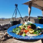 Delicious Greek Pita Breakfast Sandwiches on the Blackstone by CampingForFoodies features a blue camping plate with the open-faced sandwich on it. The plate is set on a campfire log. There is a cast iron Dutch oven and a Blackstone grill in the background with distant mountain views.