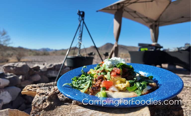 Delicious Greek Pita Breakfast Sandwiches on the Blackstone by CampingForFoodies features a blue camping plate with the open-faced sandwich on it. The plate is set on a campfire log. There is a cast iron Dutch oven and a Blackstone grill in the background with distant mountain views.