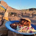 Shredded BBQ Beef Sandwiches by CampingForFoodies features a blue camping plate set on a campfire log. The plate is filled with a BBQ beef sandwich, baked beans and coleslaw tossed in a creamy dressing.