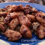 Blackstone Bang Bang Chicken by CampingForFoodies features a blue camping plate set on a campfire ring. The plate is filled with a bed of rice that is topped with golden-brown pieces of glazed chicken.