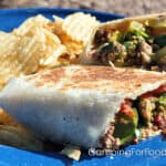Blackstone Cheesy Bacon-Burger Toasted Wraps by CampingForFoodies features a blue camping plate holding a golden brown toasted burrito-style folded tortilla that is stuffed with a hamburger and bacon mixture, green spinach, melted cheese and ripe tomatoes. Potato chips are piled next to the sandwich.