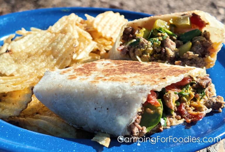 Blackstone Cheesy Bacon-Burger Toasted Wraps by CampingForFoodies features a blue camping plate holding a golden brown toasted burrito-style folded tortilla that is stuffed with a hamburger and bacon mixture, green spinach, melted cheese and ripe tomatoes. Potato chips are piled next to the sandwich.