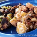 Blackstone Cheesy Bacon Chicken by CampingForFoodies features a blue camping plate filled with cubed chicken covered in melting cheese and tossed in cubed potatoes and golden brown bacon. Charred Brussels sprouts and on the side.