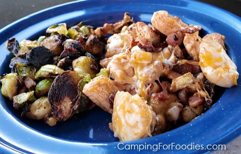 Blackstone Cheesy Bacon Chicken by CampingForFoodies features a blue camping plate filled with cubed chicken covered in melting cheese and tossed in cubed potatoes and golden brown bacon. Charred Brussels sprouts and on the side.