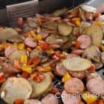 Blackstone Cowboy Stir-Fry by CampingForFoodies features a griddle filled with sliced sausage, potatoes, onions, red and yellow bell peppers. Two spatulas are being used to toss the food as it cooks on a flattop Blackstone griddle.