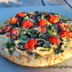 Alfredo Pizza by CampingForFoodies features a pizza on a cutting board that is set on a picnic table. The crust is topped with red tomatoes, green basil strips, white alfredo sauce and chicken.
