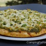 Pesto Pizza by CampingForFoodies features a pizza crust topped with green basil pesto, grilled chicken, white cheese and torn green basil leaves. The pizza is on a pan set on a small table outdoors.