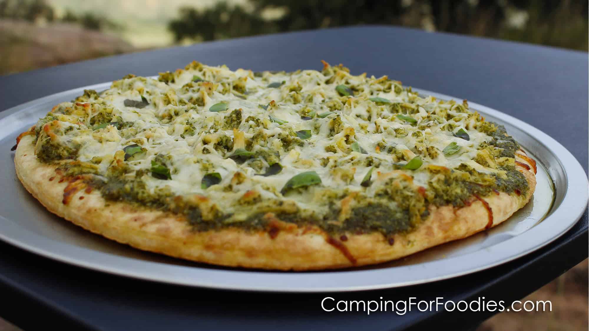 Pesto Pizza by CampingForFoodies features a pizza crust topped with green basil pesto, grilled chicken, white cheese and torn green basil leaves. The pizza is on a pan set on a small table outdoors.